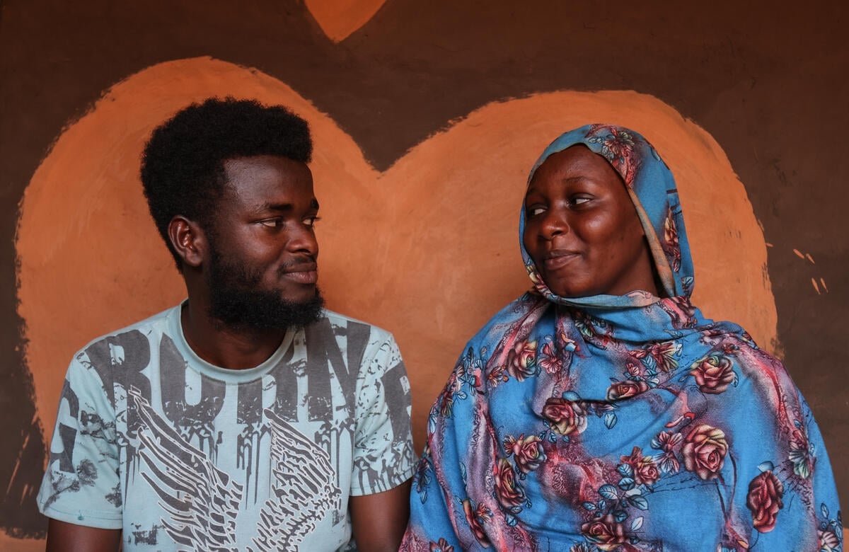 A woman and a man sit together, smiling at each other, in front of a heart-shaped wall painting