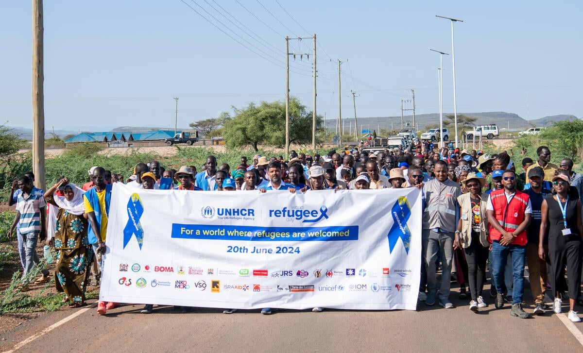 A large group of people walk holding a huge banner which reads 'World Refugee Day: For a world where refugees are welcomed. 20th June 2024'