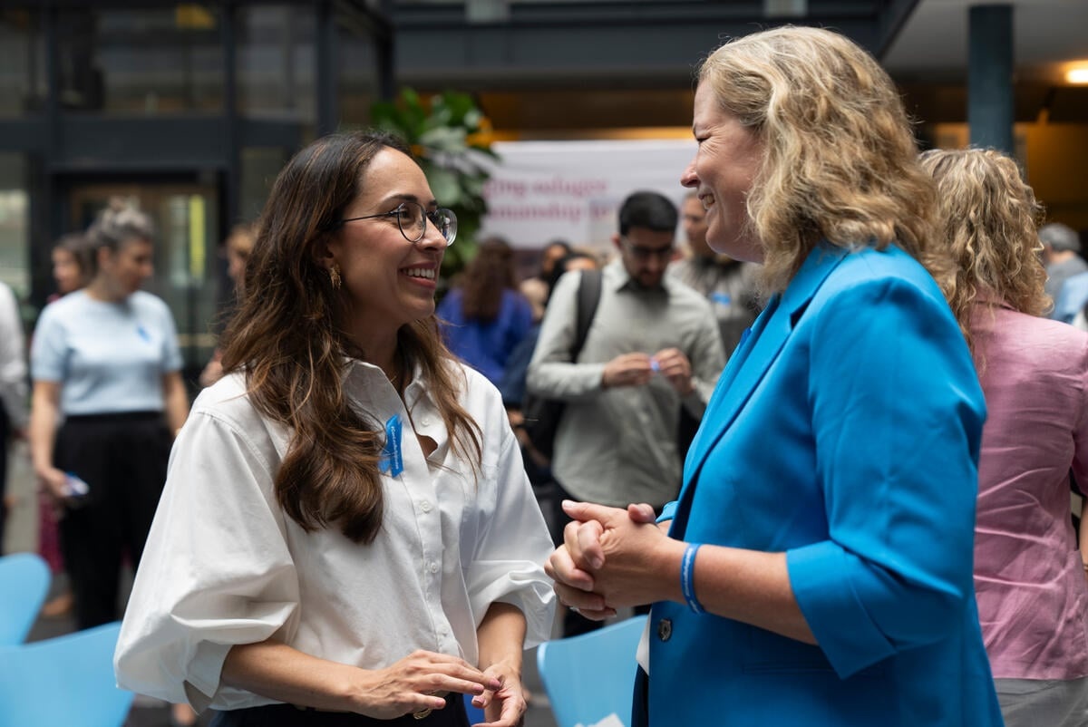 Two smiling women speak together, with a crowd in the background. They are both wearing #WithRefugees blue ribbons.