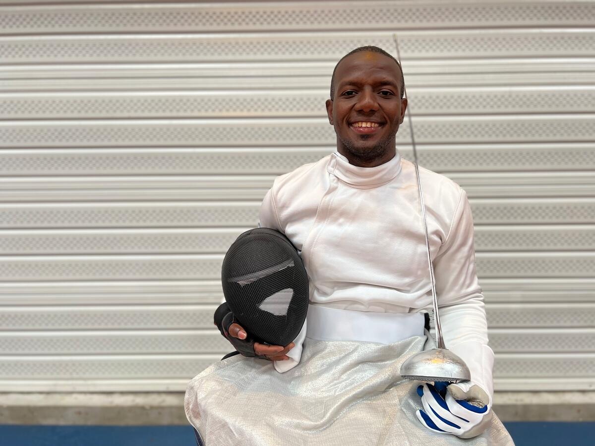 A man wearing a white fencing jacket poses with a fencing blade and mask.