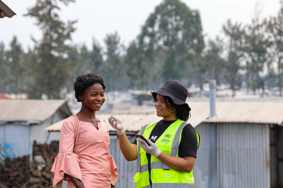 A woman in a high-vis UNHCR-branded vest and holding a phone talks to another woman in a camp.