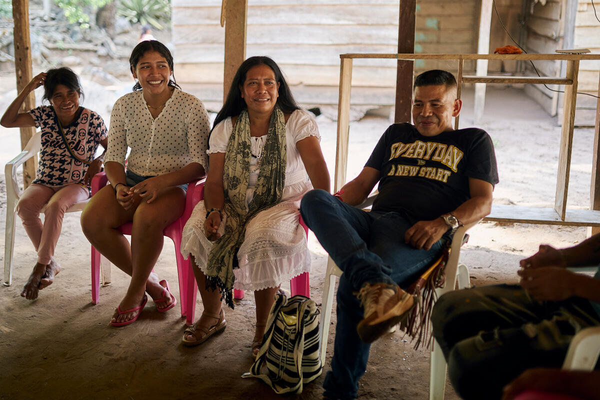 A man and three women sit on plastic chairs under an open-sided shelter.