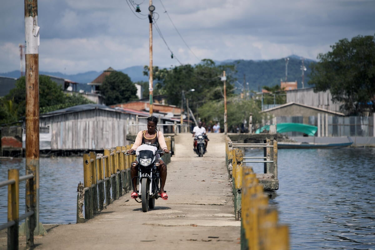 A woman woman drives a motorcycle across a walkway over a bay.