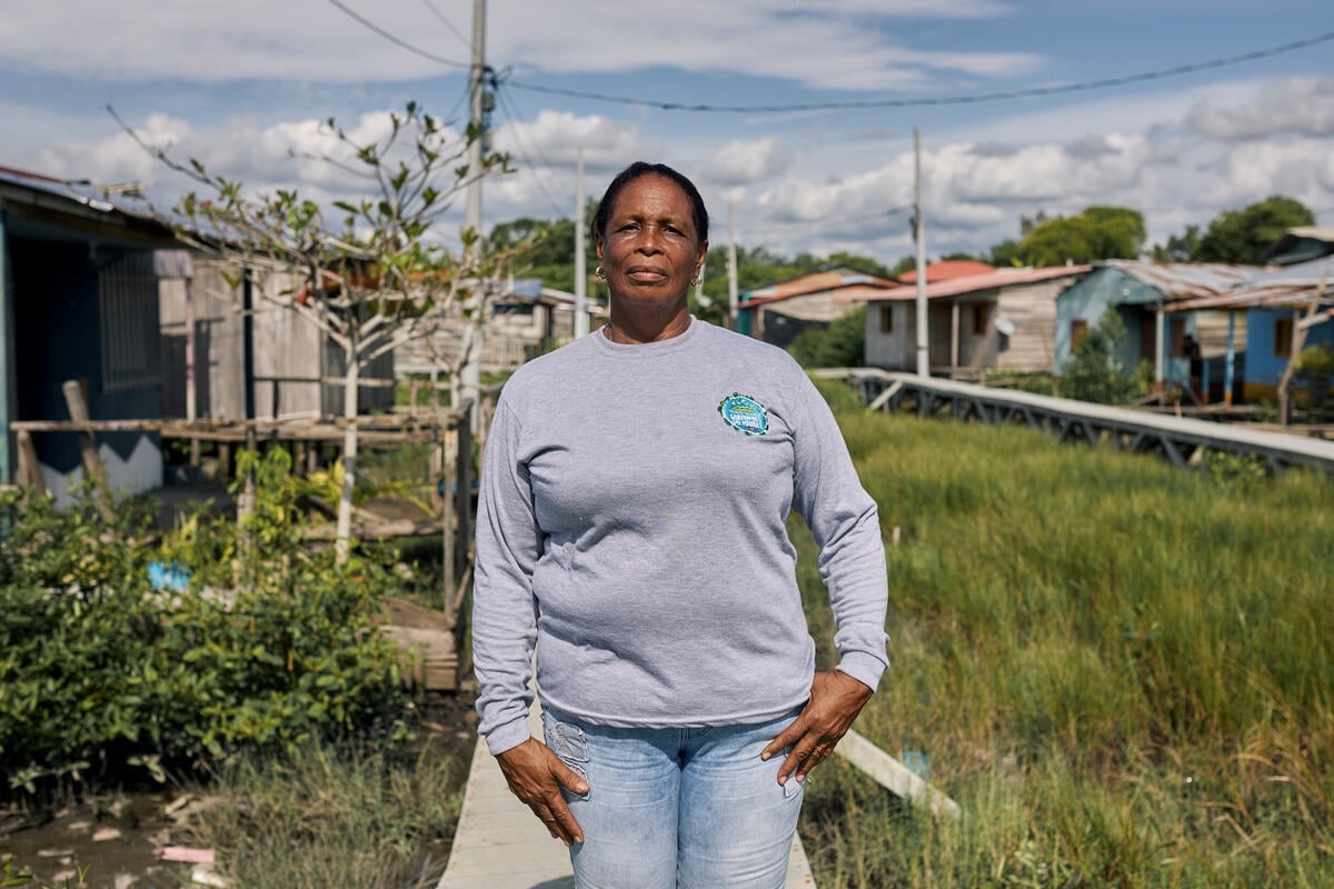 A woman in a grey sweatshirt stands on a boardwalk over marshland surrounded by wooden shacks.