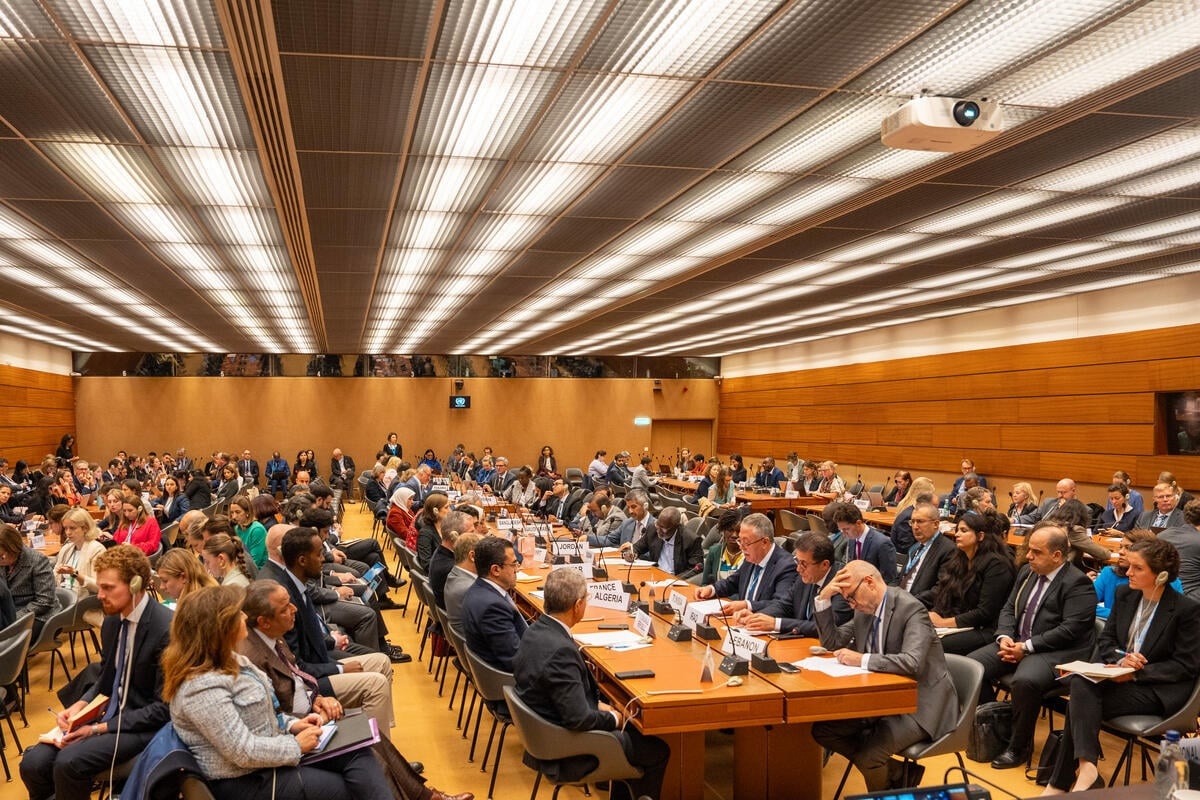 A packed audience sits in rows around long tables running the length of a brightly lit room