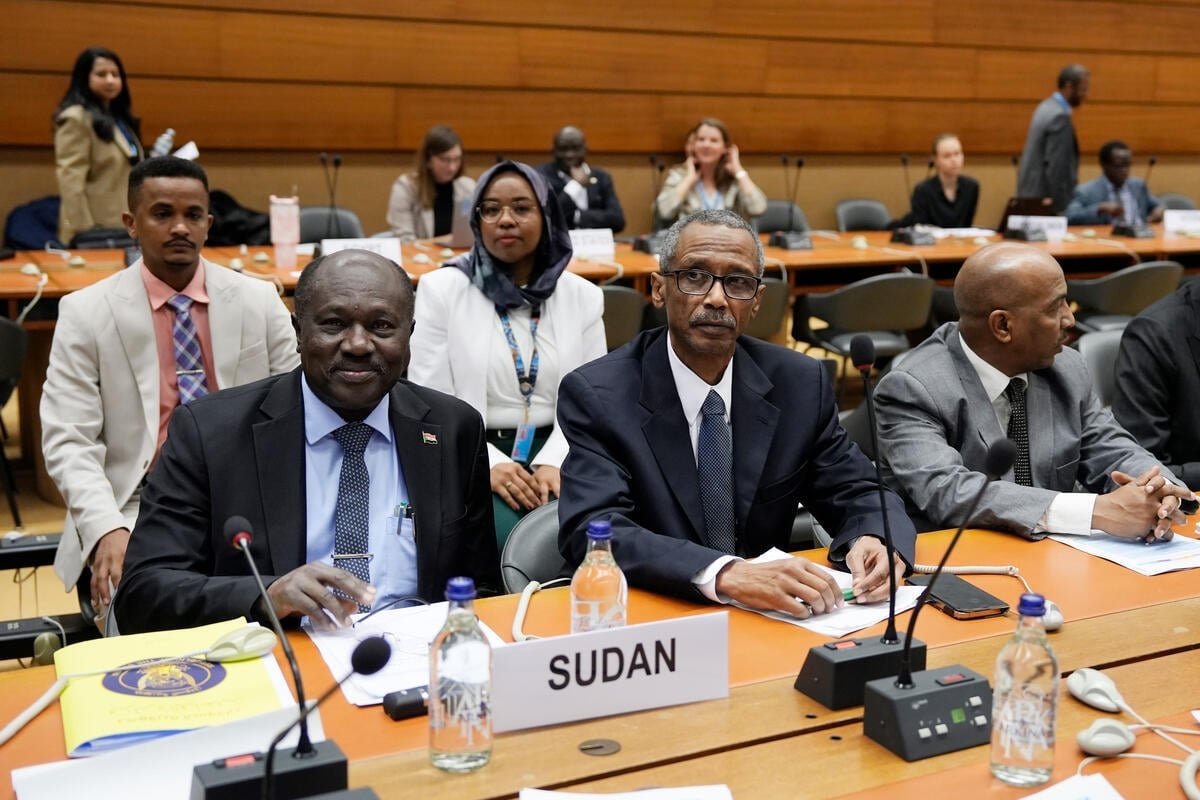 Two men in suits sit at a table in front of microphones and a sign reading 'Sudan'