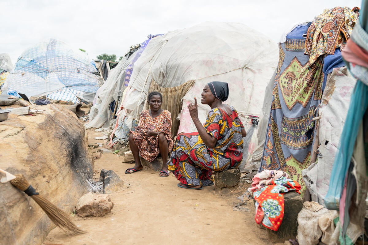 A woman sits talking to another woman in front of a makeshift shelter.