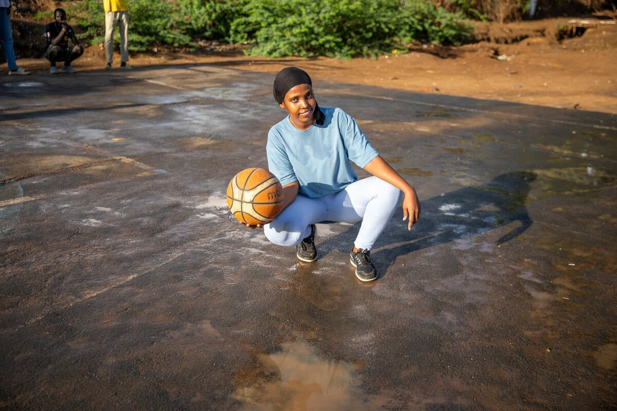 A young woman squats down on a basketball court holding a basketball.
