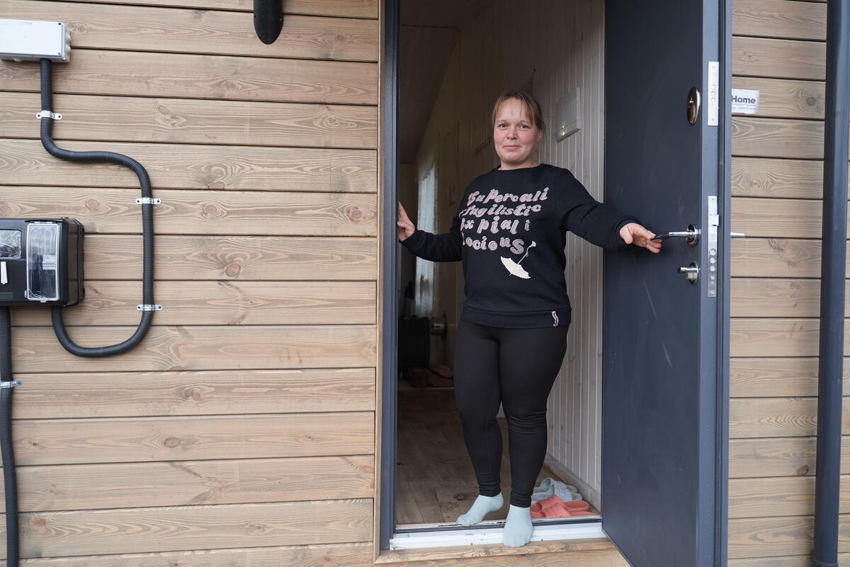 A woman stands in the open doorway of a wooden-clad prefabricated house