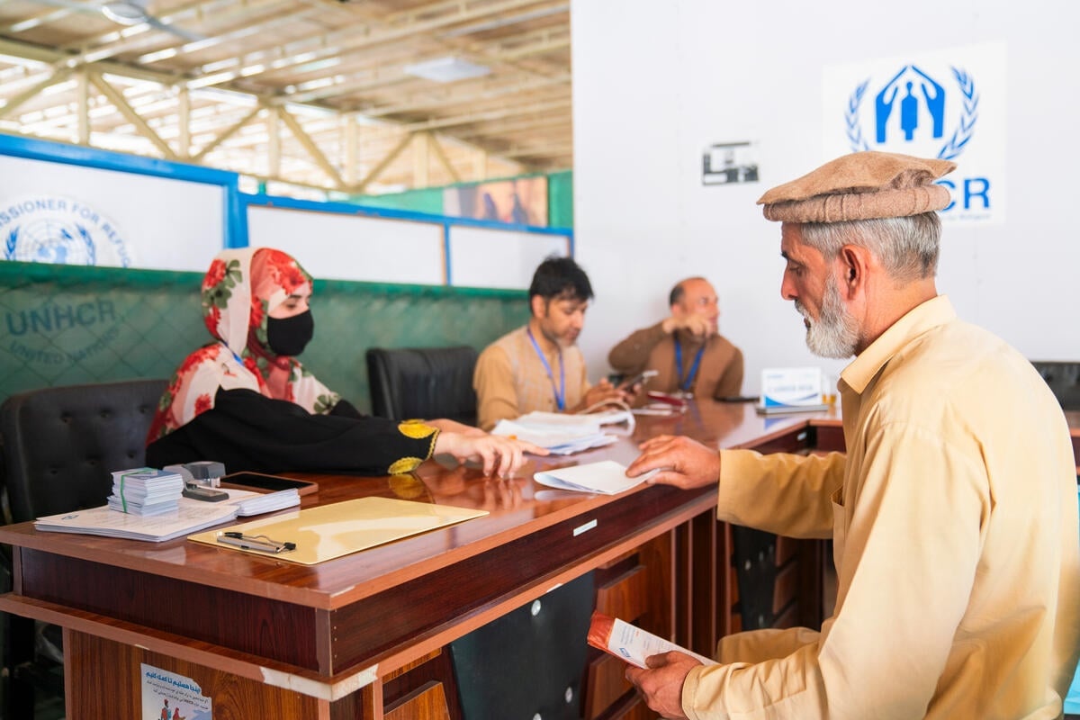A man wearing a traditional pakol or rolled-up cap receives a document from a woman sitting behind a desk with other colleagues with UNHCR logos on the walls behind her.