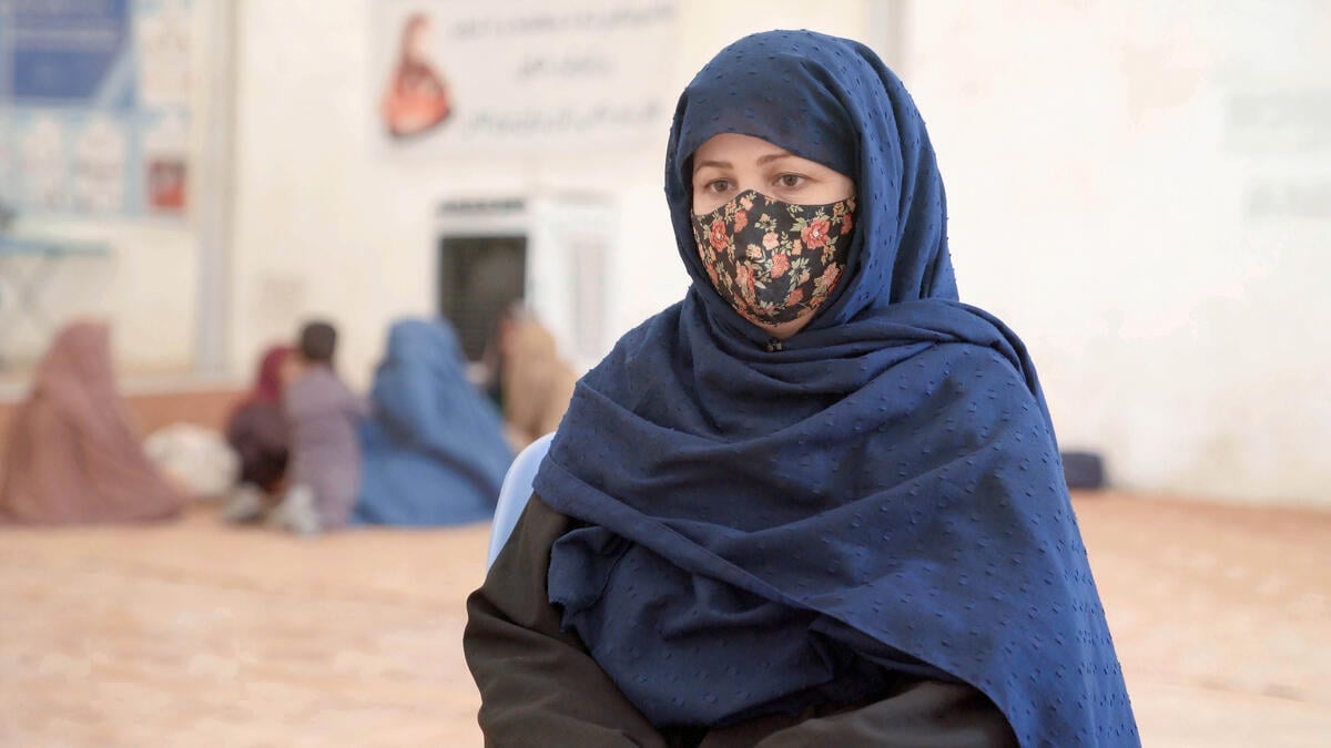A woman wearing a blue headscarf and floral facemask sits on a blue plastic chair inside a large rub hall tent.