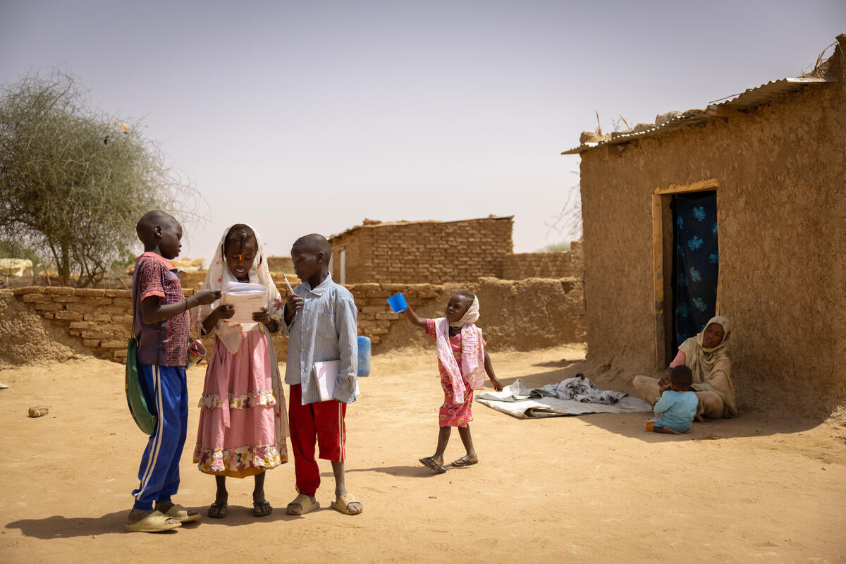 Four children stand together with their mother and younger sibling sitting in the shade outside a small mud-brick house.  