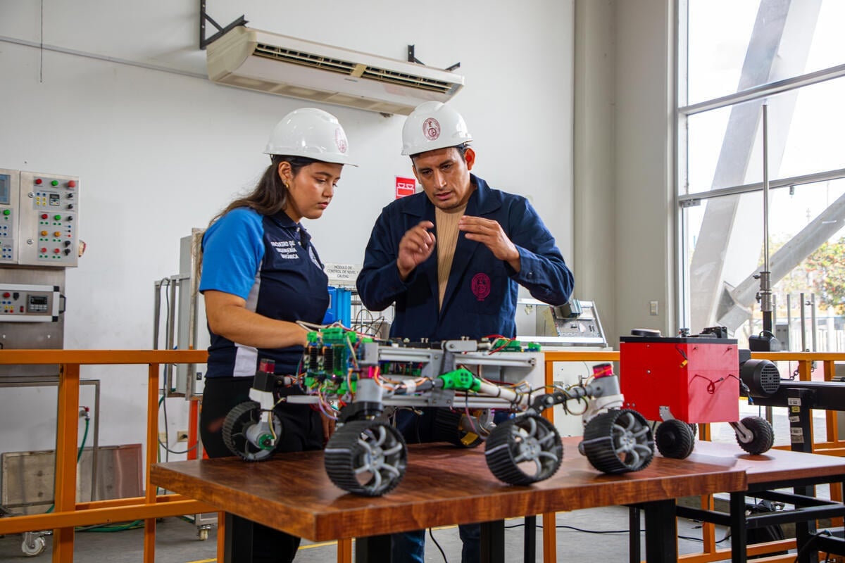 A woman and a man wearing white safety hats stand behind a six-wheeled robot on a bench in a laboratory 