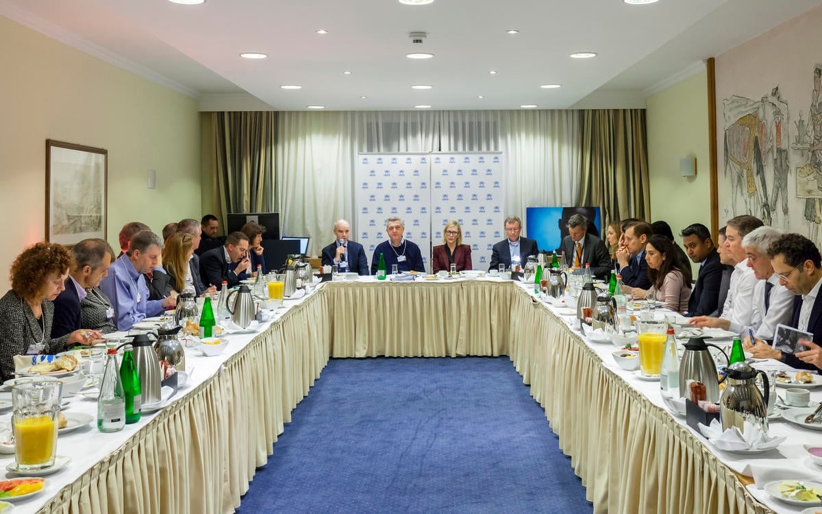 A large group of people seated indoors around a U-shaped table covered in food and drinks