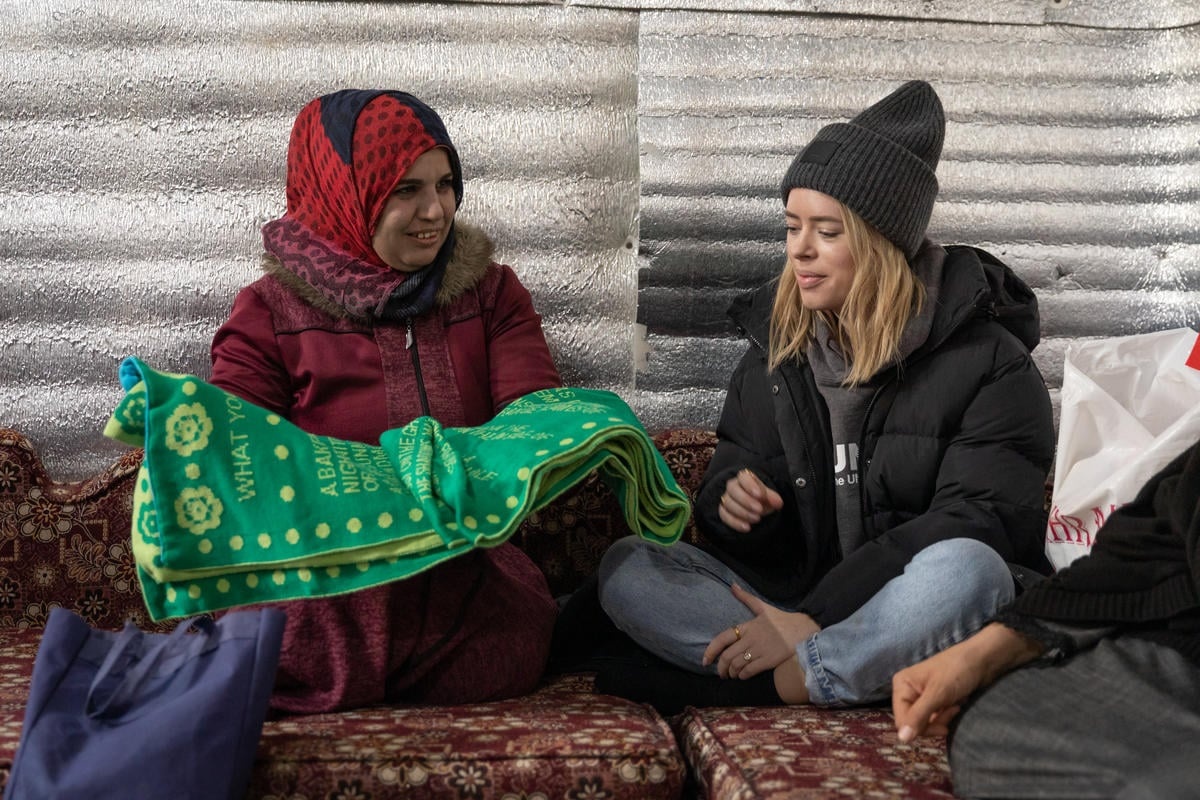 Tanya Burr sits on the ground, next to a refugee woman showing her a crocheted scarf.