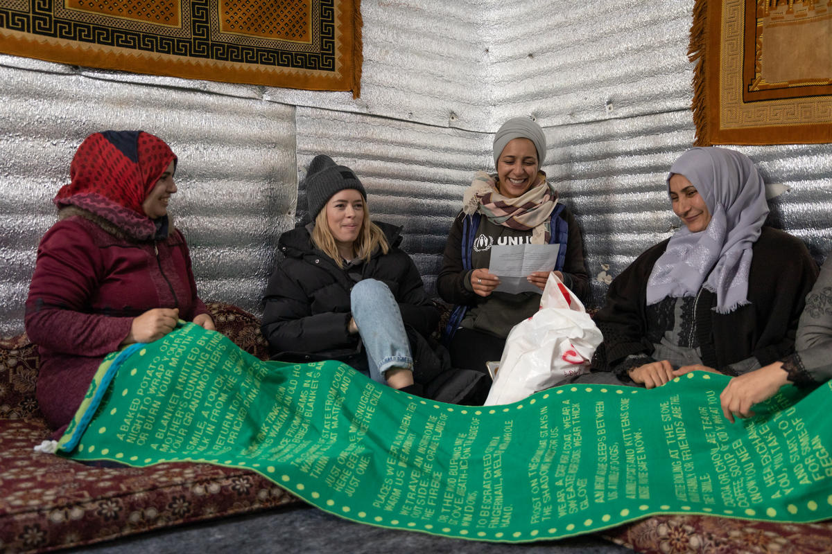 Tanya Burr sits on the ground with three refugee women. They hold a big green scarf in their hands.