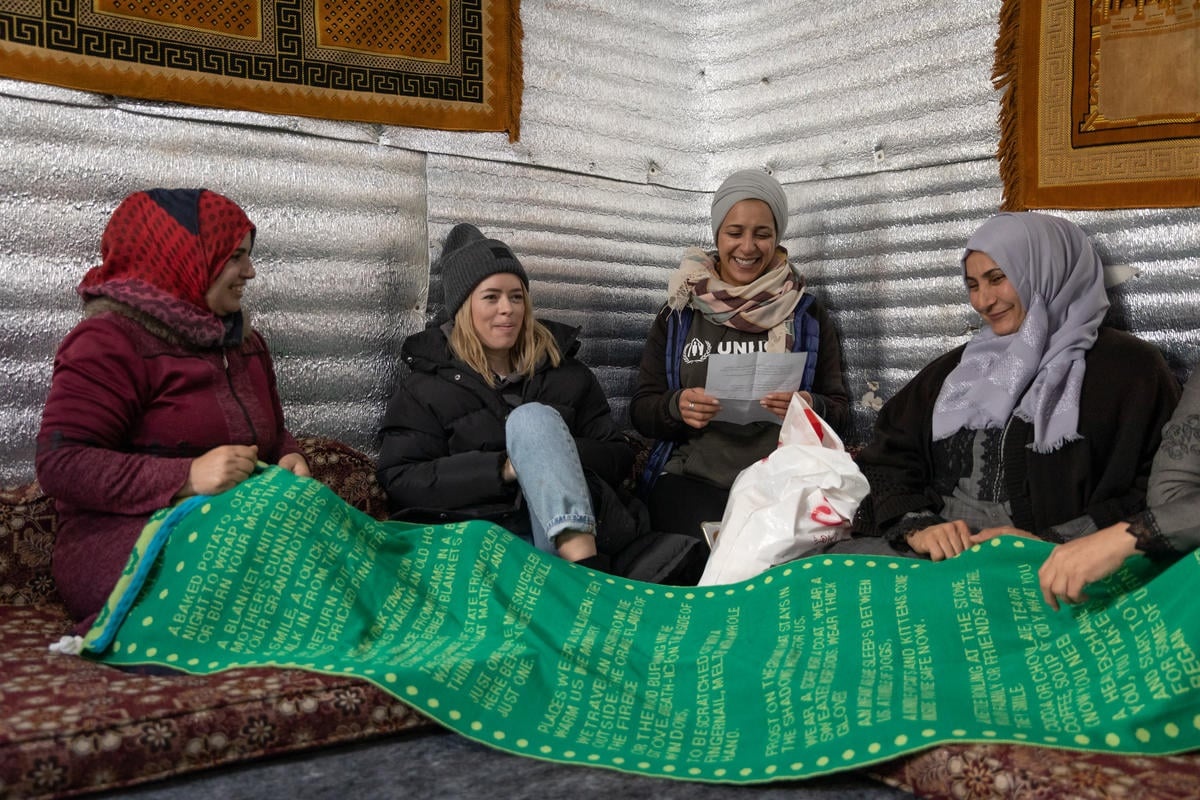 Tanya Burr sits on the ground with three refugee women. They hold a big green scarf in their hands.