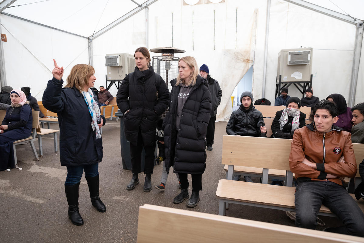 Tanya Burr stands in a waiting room where people sit on benches around her.