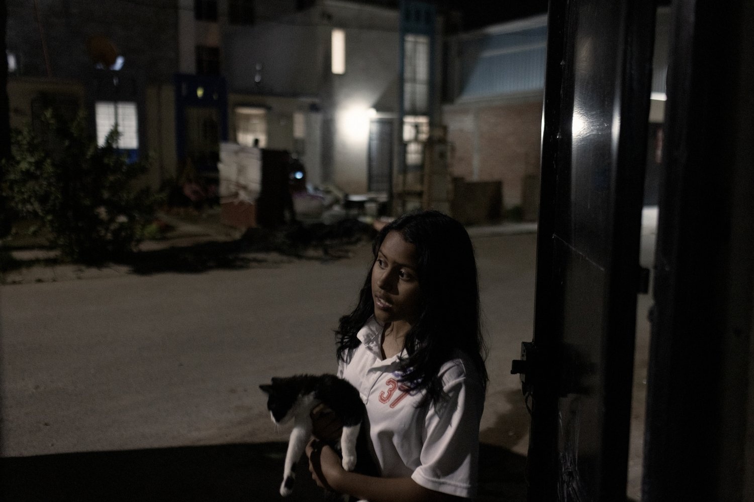 A girl stands by a gate in a dark street holding a cat.