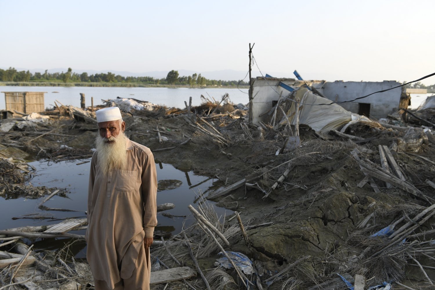 An elderly man stands in front of a house destroyed by flooding.