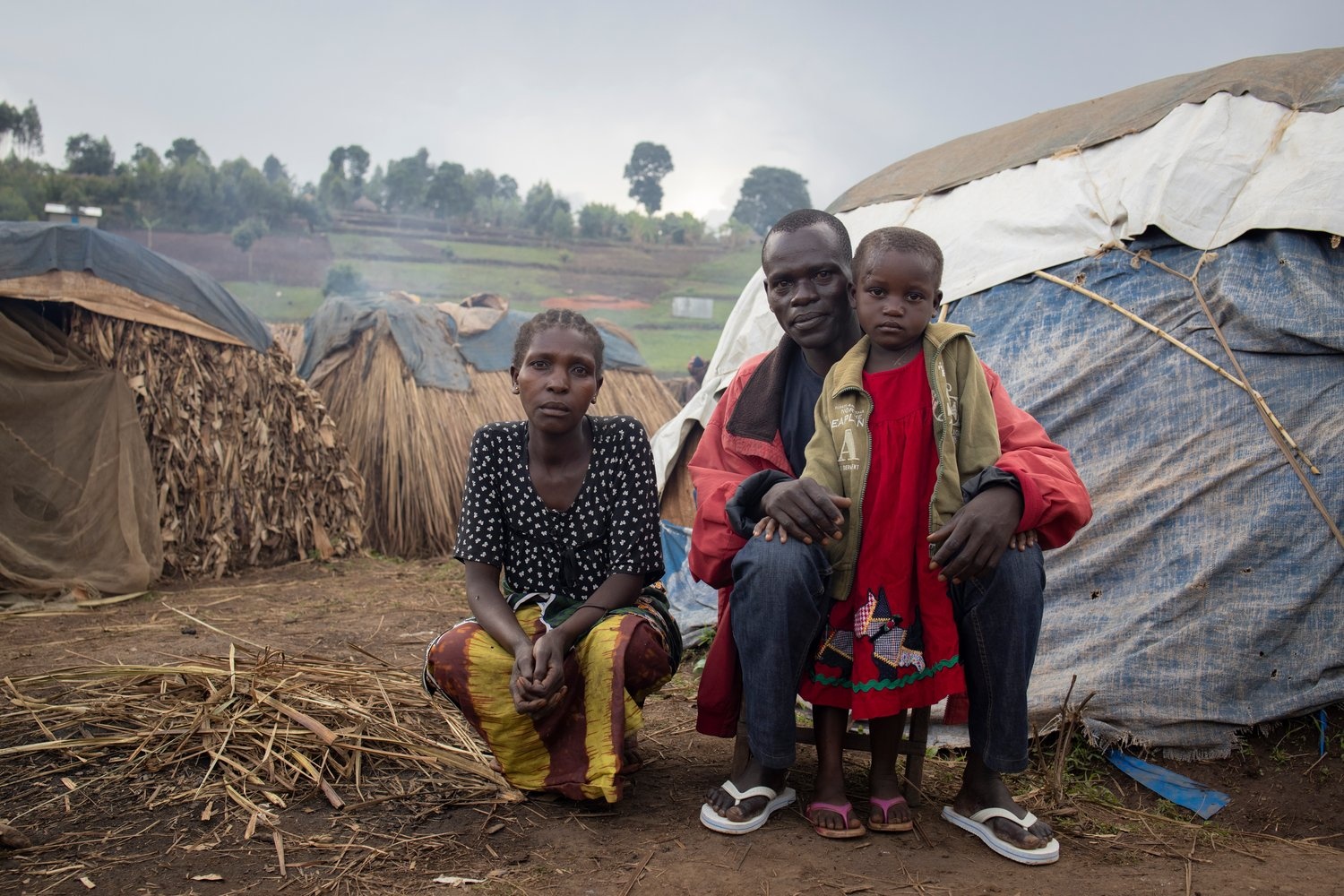 A small girls stands with her parents in front of a basic shelter.