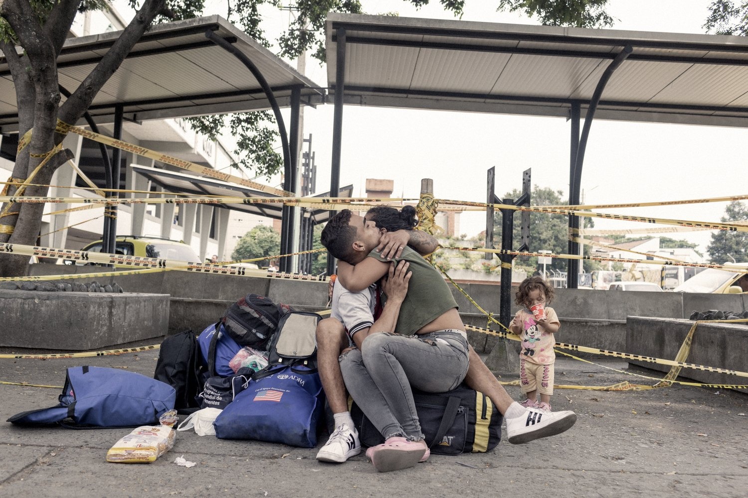 A woman hugs a man sitting on luggage while a small girl drinks from a disposable cup.