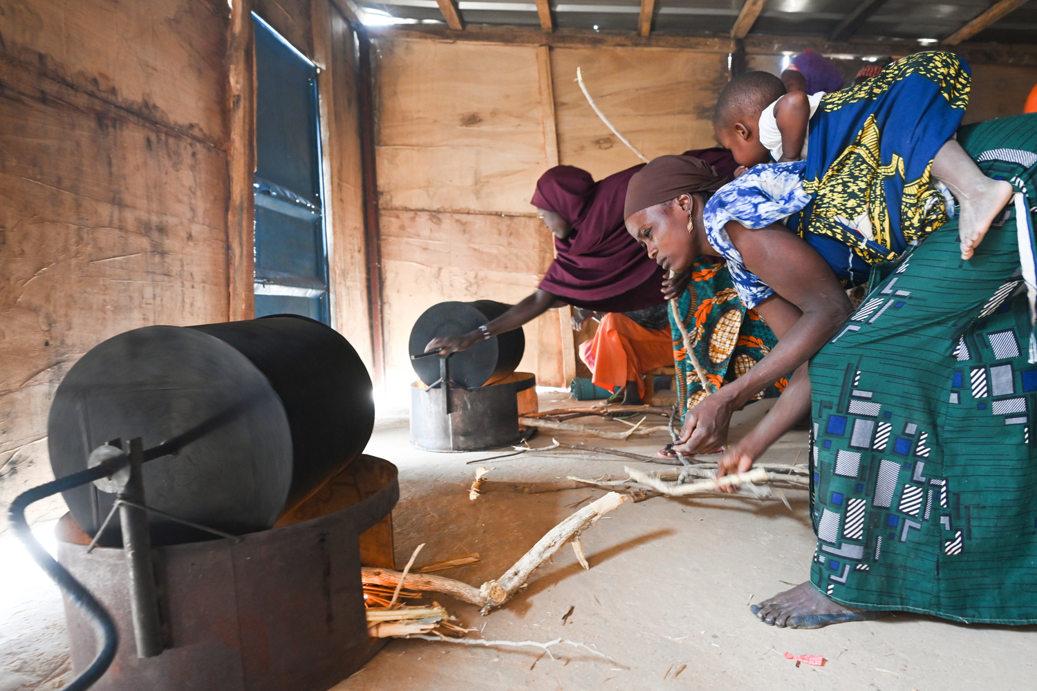 Two women stoke two wood-burning stoves in a shed-like structure.
