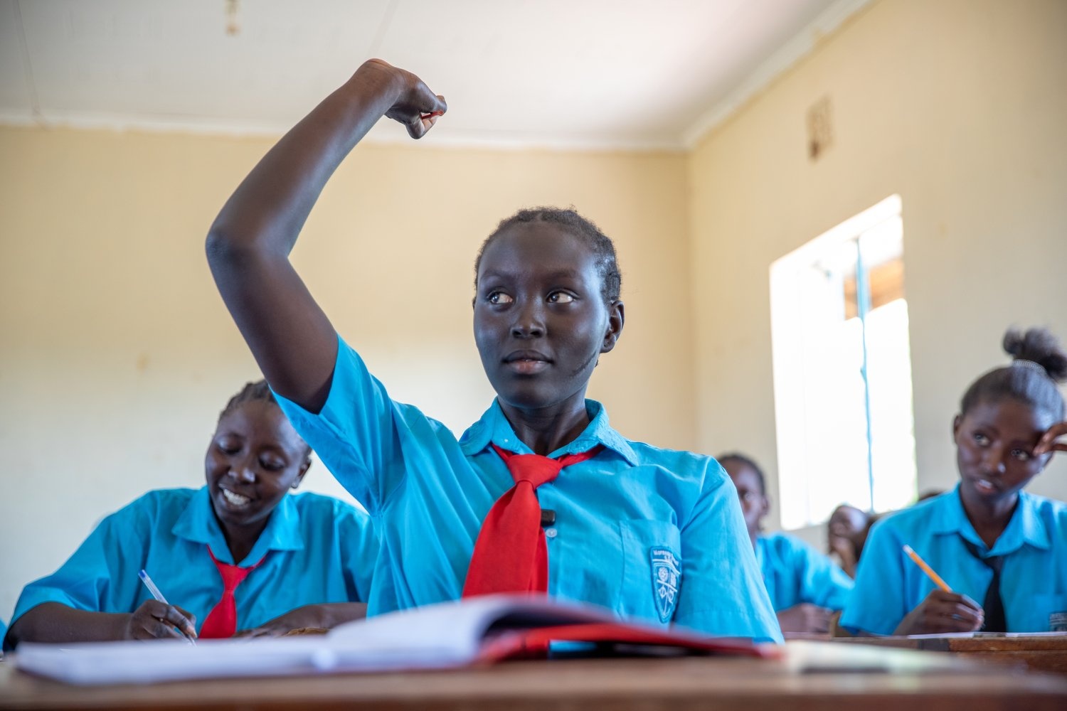 A teenage girl in a school uniform raises her hand in a classroom.