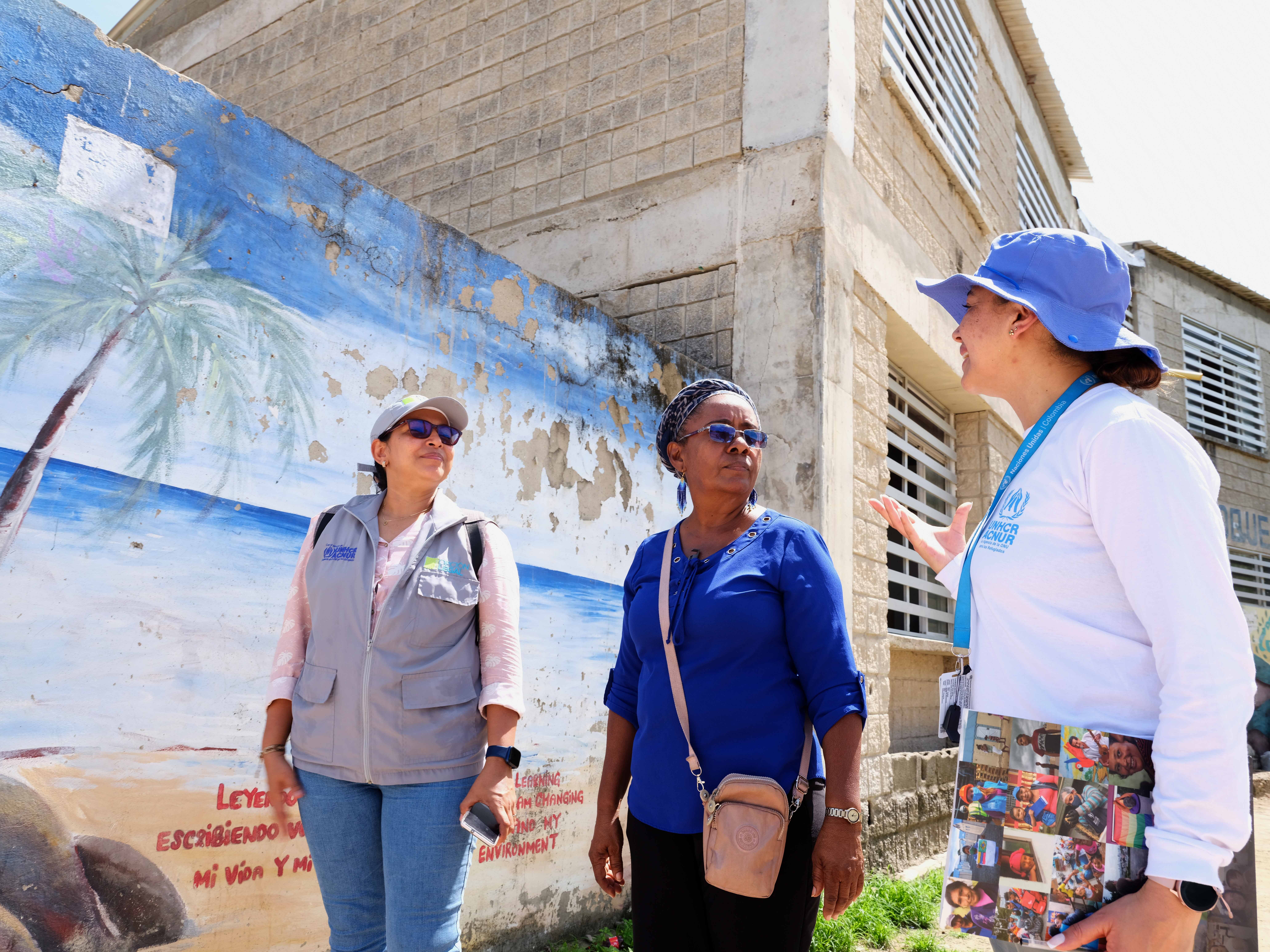two UNHCR staff walk with a woman outside concrete buildings