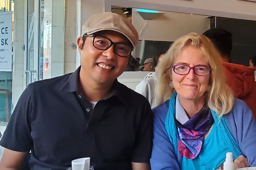 a man in a navy polo shirt and beige newsboy cap sits to the left of a blond woman with shoulder length hair and dark rimmed glasses