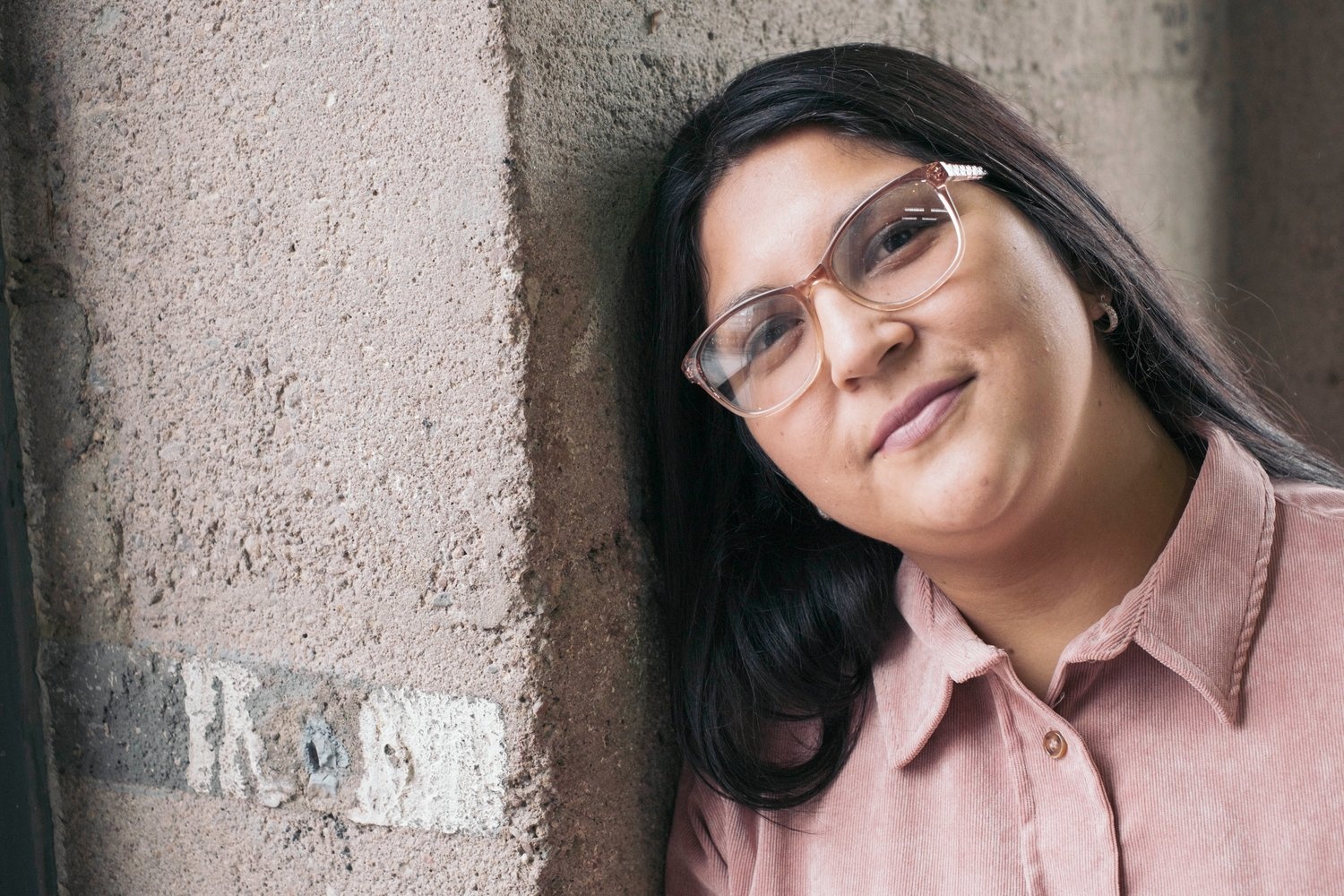 A young woman in a pink shirt leans her head against a wall.