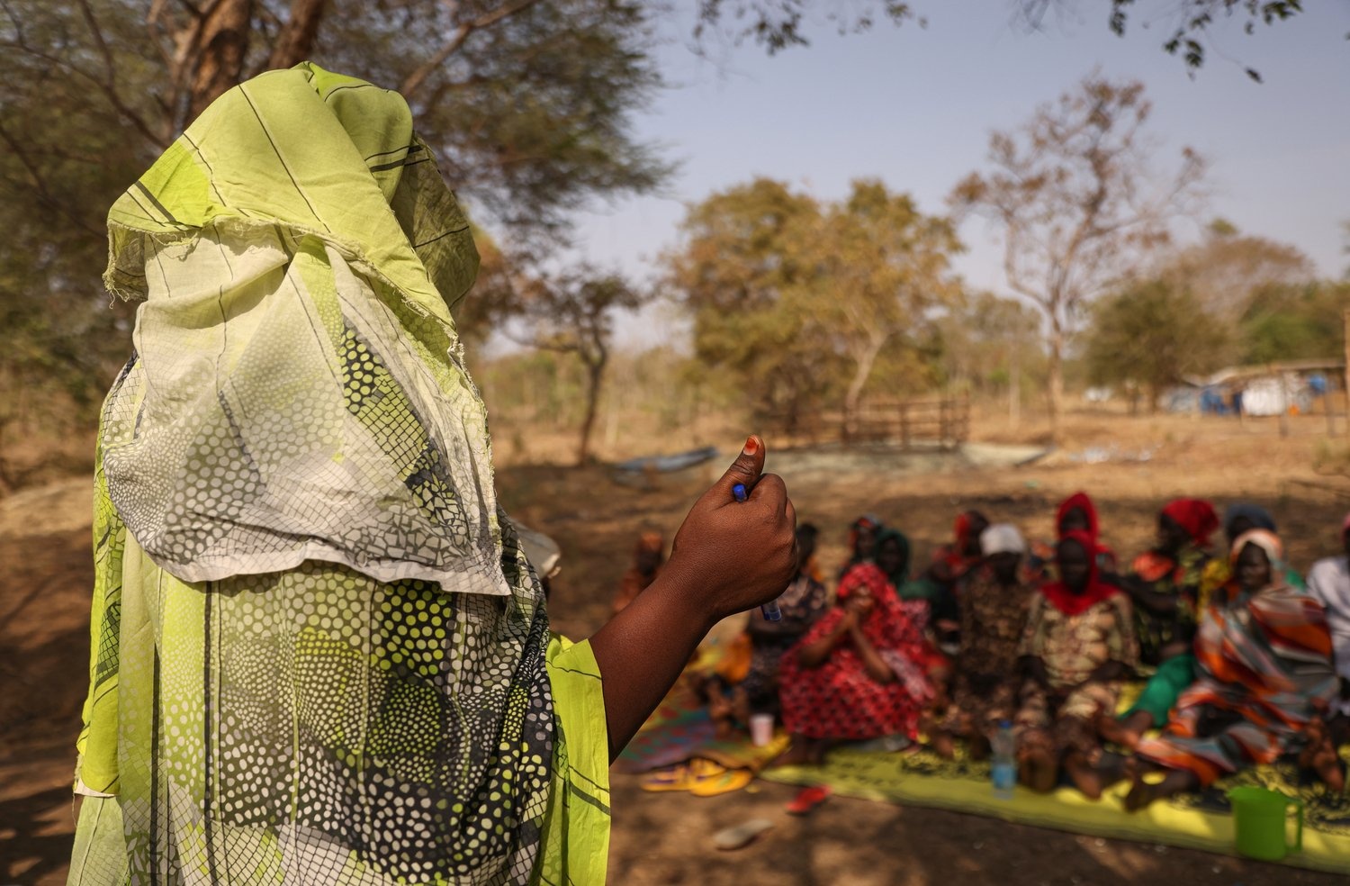 A woman speaks in front of a group seated outside in the shade of a tree.
