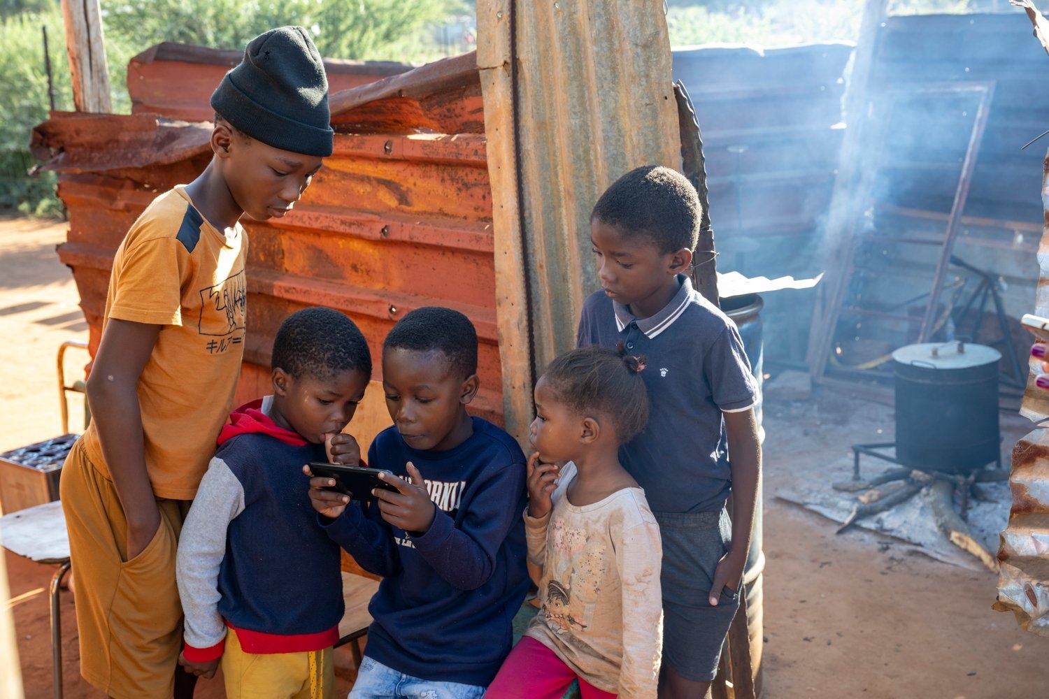 A young boy holds a phone and stares at the screen along with four other children. 