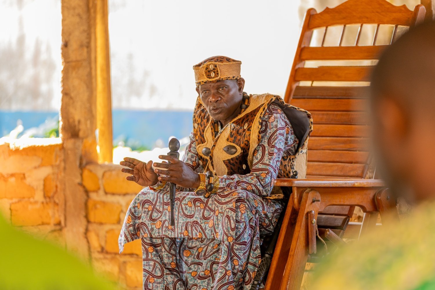 A man in colourful robes sits in a large wooden chair and speaks 