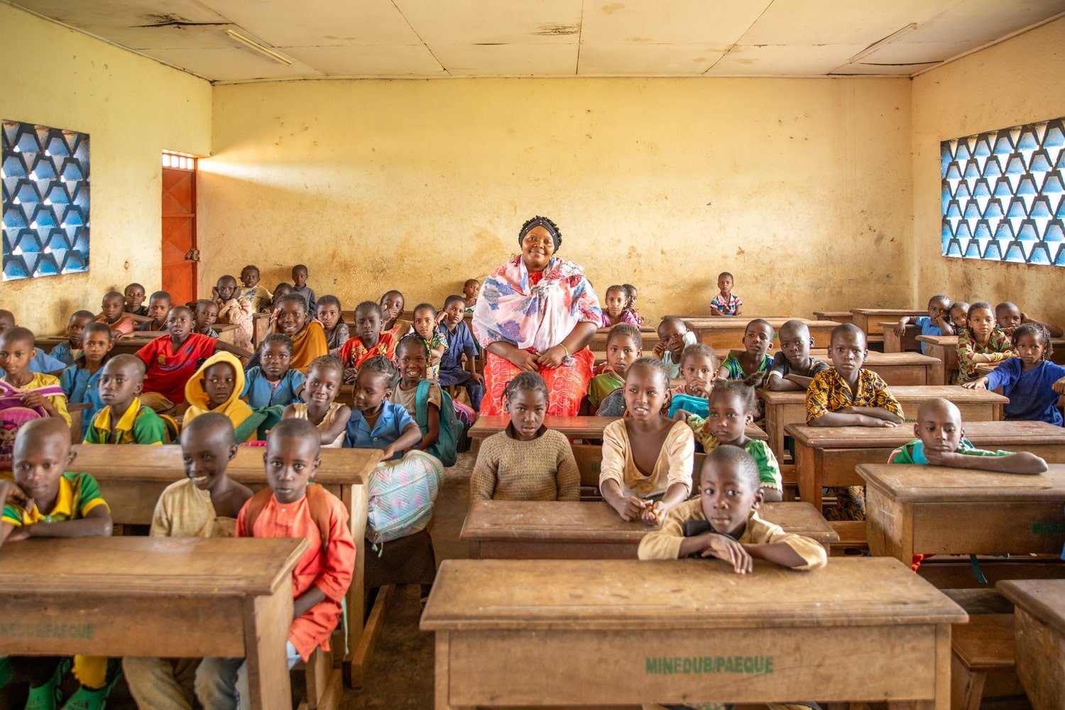 A woman stands in a classroom surrounded by pupils sitting at wooden desks