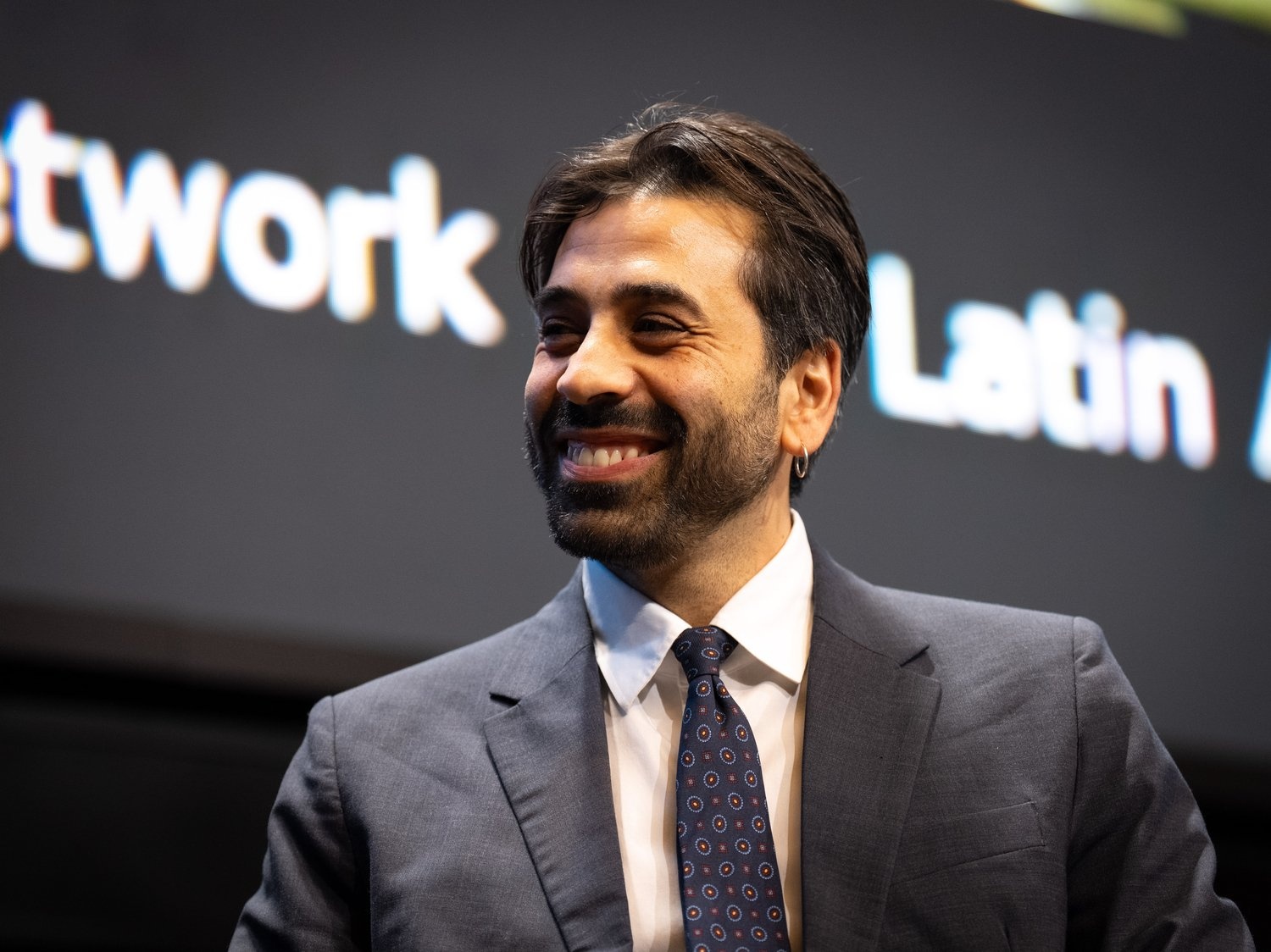 A man in a suit and tie smiles in front of a large screen displaying text