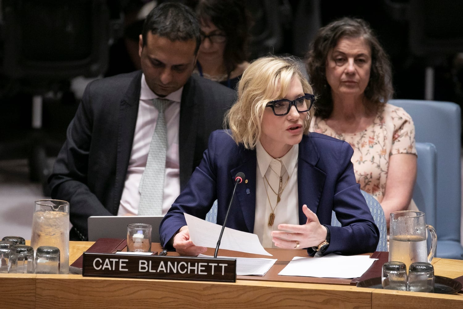 Cate Blanchett speaks into a microphone from behind a desk and gestures with one hand while holding a speech in the other