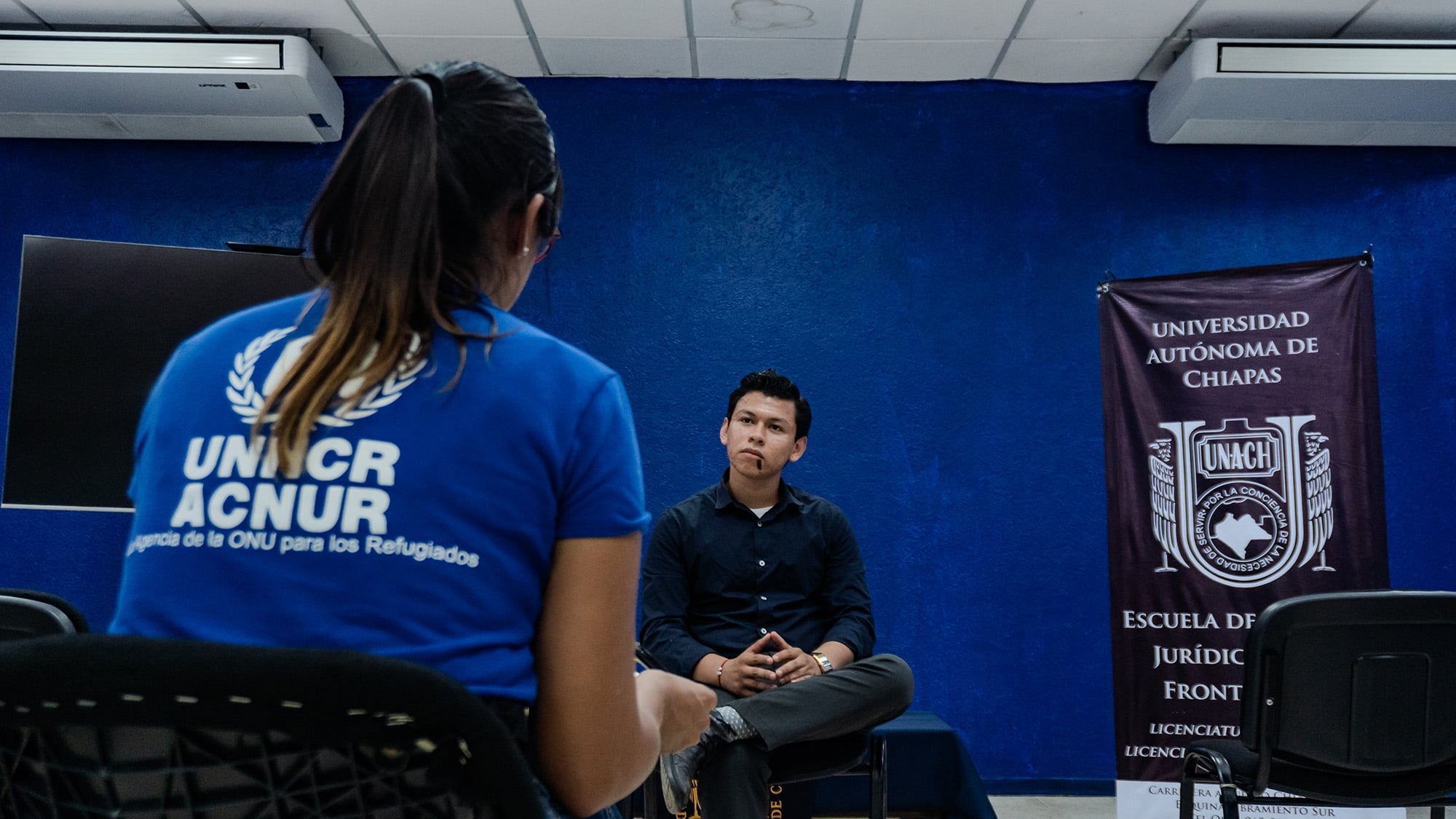 With her back to the camera, a woman wearing a blue T-shirt with a white UNHCR logo is talking to a young man dressed in black
