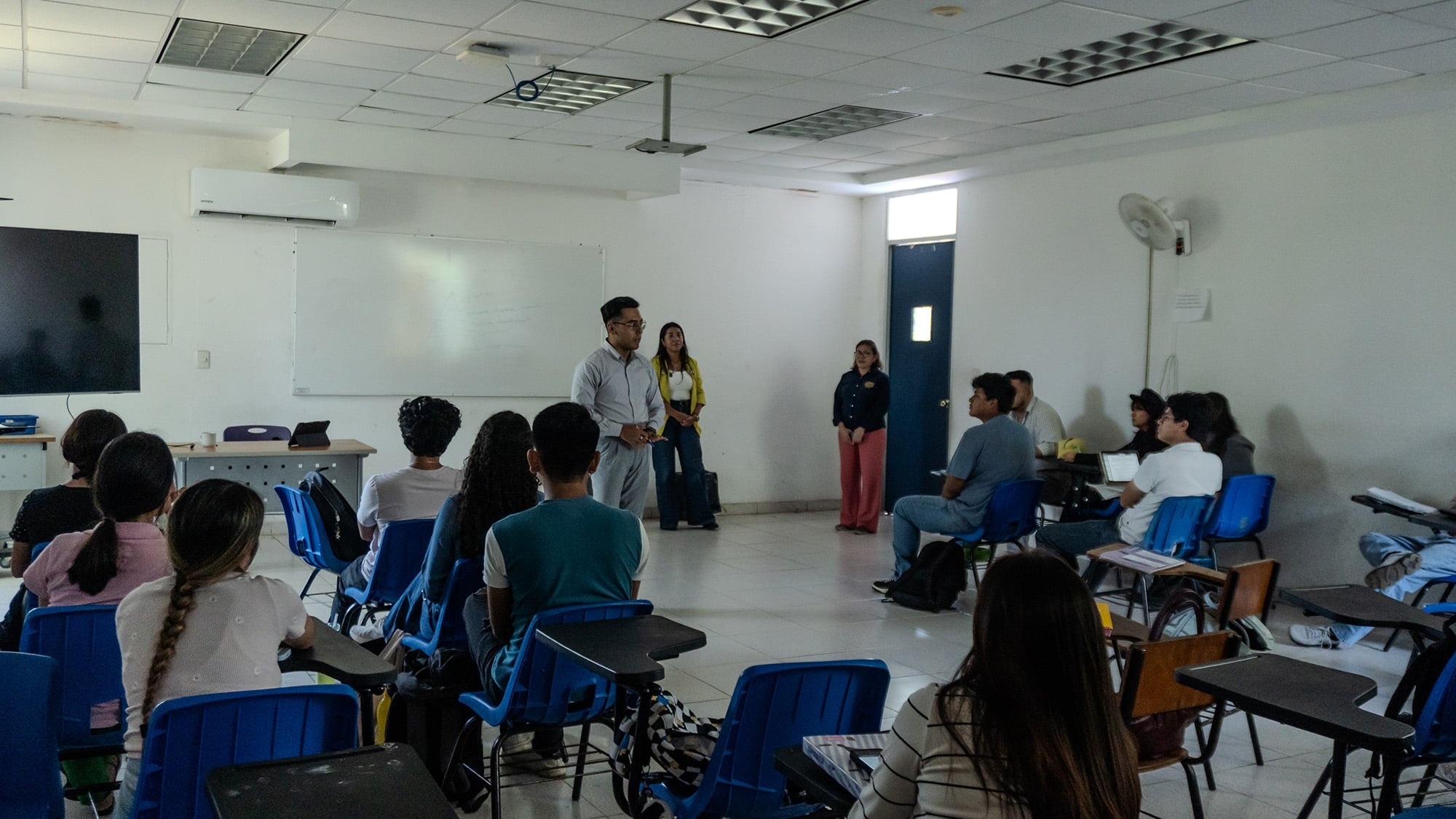 At least fifteen young people, seated at desks in a classroom, are listening attentively to a man speaking to the group. Behind the man, two other women are standing