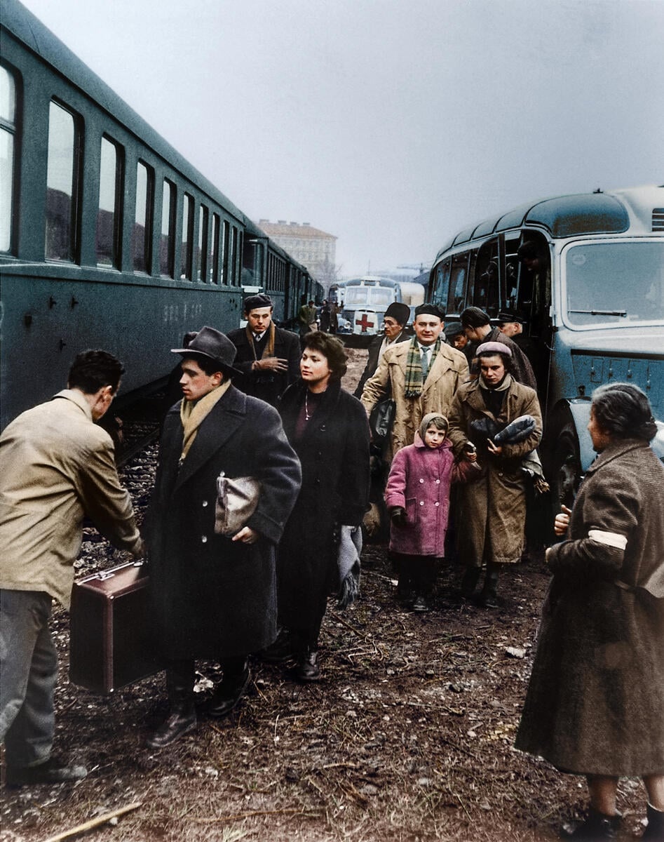 Men, women and children wearing warm, 1950s clothes carry suitcases from buses to a waiting train