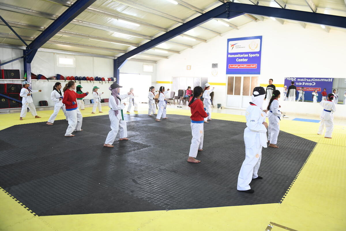 A group of girls wearing white uniforms practice Taekwondo moves inside a large hall.