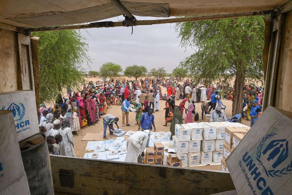 A view from the back of a truck showing workers unloading relief supplies for refugees waiting nearby.