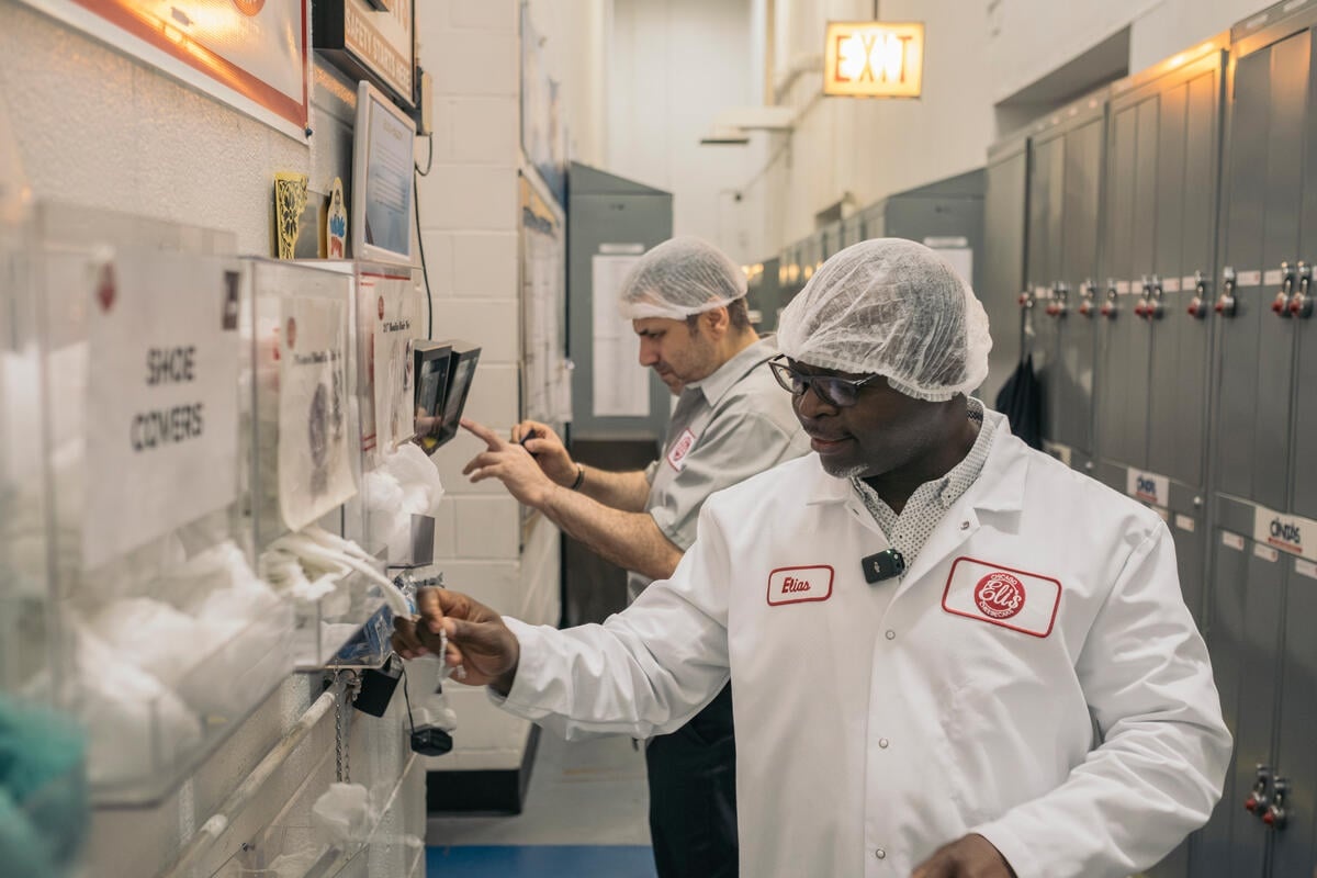 A man wearing a white coat and hairnet stands in the locker room of a factory.