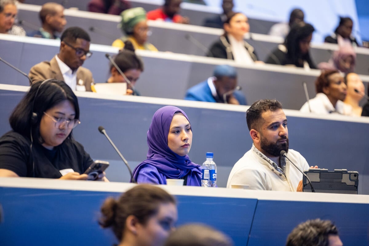 Participants at an event sit behind rows of desks in a conference centre.