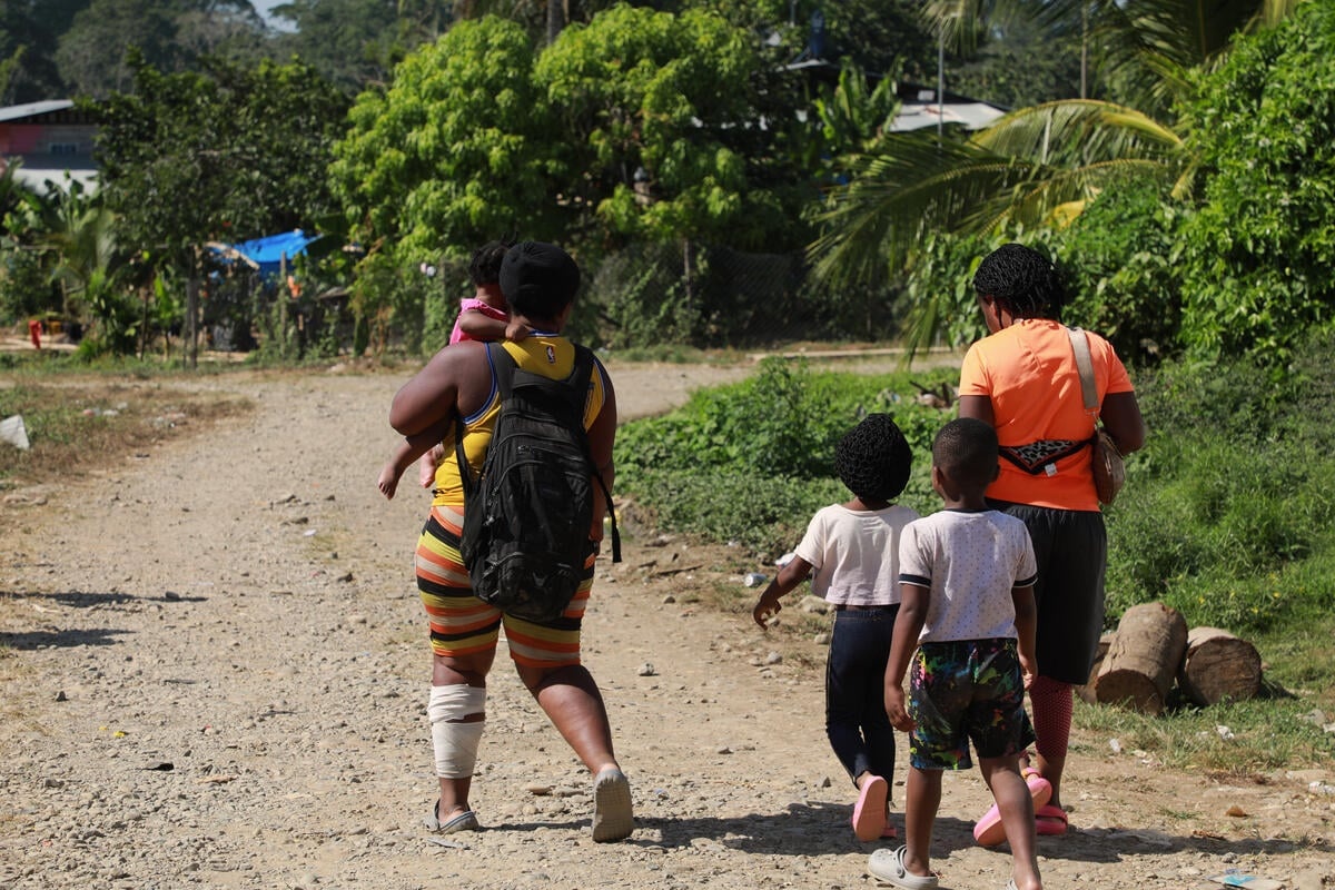 A woman with a bandage on her leg carries a toddler while two small children walk alongside another woman.