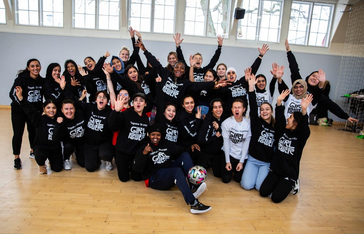 A group of around 30 young women in Girl Power sweaters smile with hands in the air