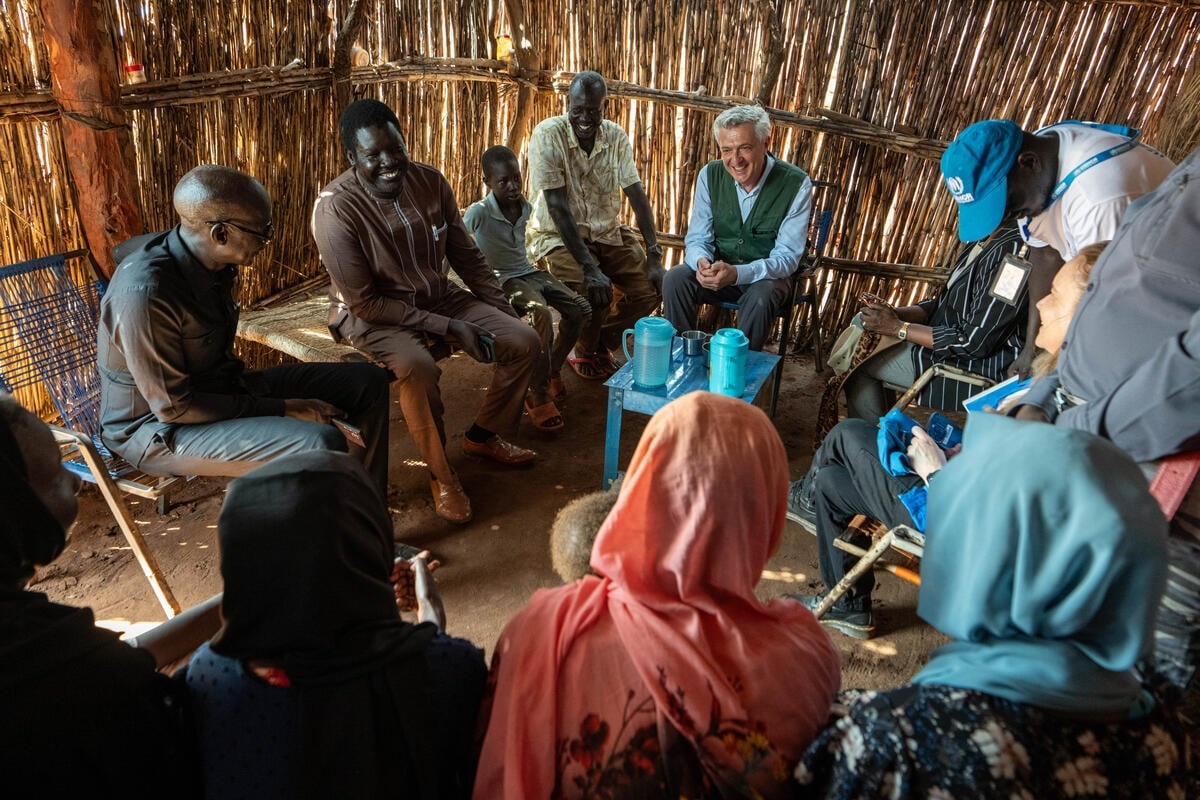 A group of people sit in a circle of chairs inside a structure with wooden thatched walls