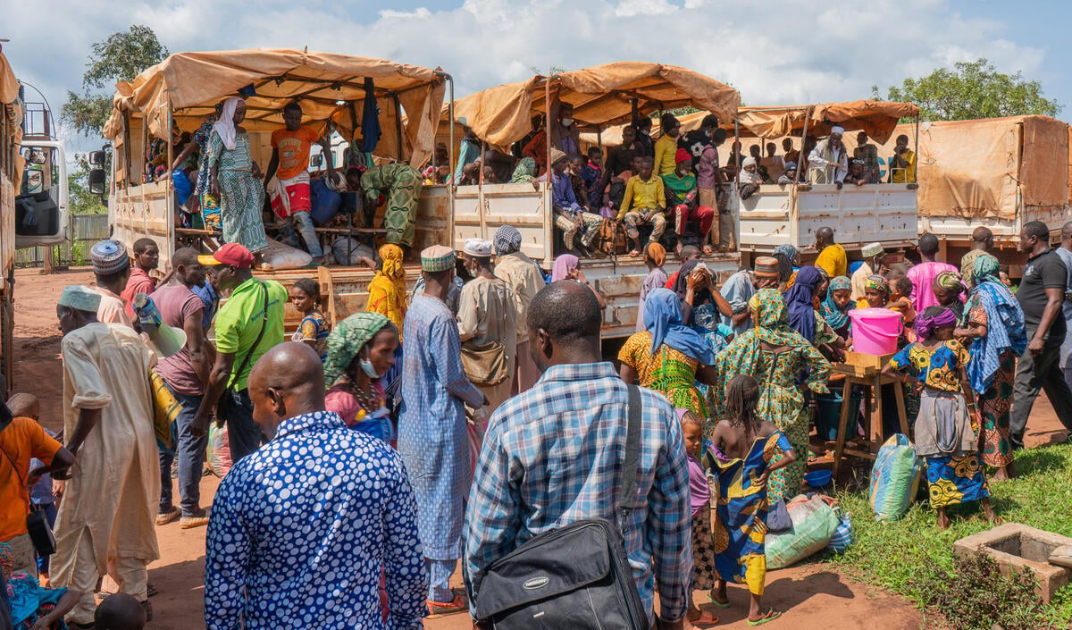 A crowd of people gathers behind several trucks filled with passengers