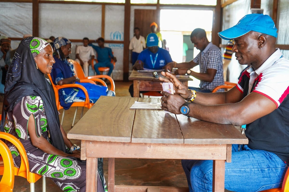 A woman sits across a desk from a man in a blue UNHCR cap as he counts out money.
