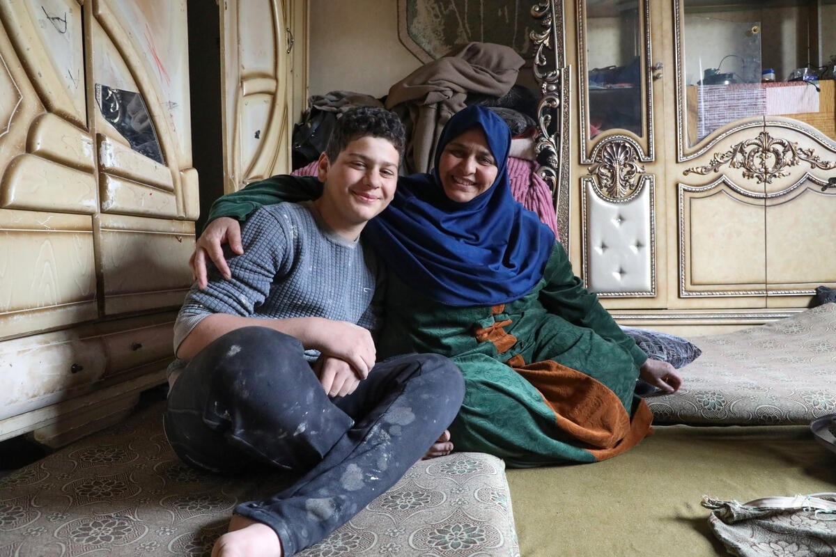 A mother has her arm round her teenage son, both sitting cross-legged on floor cushions in a living room.